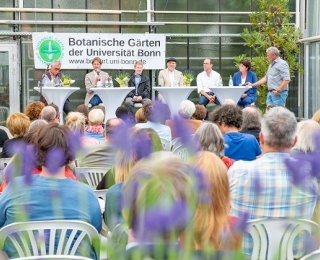 Foto einer Podiumsdiskussion auf einer Bühne in den Botanischen Gärten der Universität Bonn. Sieben Personen diskutieren an Stehtischen. Davor sitzt auf weißen Stühlen das Publikum. Im Vordergrund sind unscharf lila Blüten zu sehen.