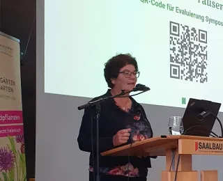 Sabine Riewenherm spricht auf einer Bühne beim Symposium des Projekts „Tausende Gärten – Tausende Arten“. Sie steht an einem Rednerpult mit dem Logo „SAALBAU“ und spricht in ein Mikrofon. Im Hintergrund ist eine Präsentationsfolie mit einem QR-Code zur Evaluation zu sehen. Links neben ihr steht ein Roll-Up mit dem Projektlogo und dem Slogan „Wildpflanzen – echt heimisch“. Es fordert zum Mitmachen auf: „Mach mit!