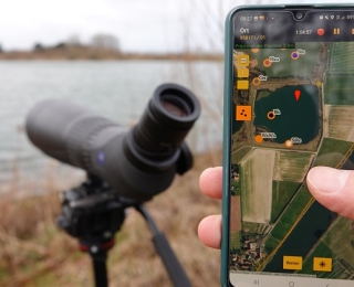 Im Vordergrund sieht man ein Handy mit geöffneter Erfassungs-App, dahinter ein Spektiv am Seeufer