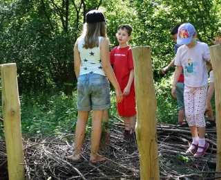 Das Foto zeigt vier spielende Kinder auf einen als Spielelement aufgeschichteten Reisighaufen in einem waldbestockten Naturerfahrungsraum.