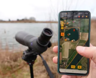 Im Vordergrund sieht man ein Handy mit geöffneter Erfassungs-App, dahinter ein Spektiv am Seeufer
