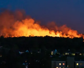 Das Foto zeigt einen großflächigen Brand in einem Waldgebiet bei Nacht. Über der Vegetation sind Flammen und Rauch zu erkennen. Im Vordergrund befinden sich Gebäude einer nahegelegenen Wohnsiedlung.