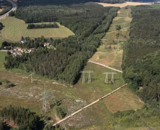 Luftaufnahme einer Wald- und Feldlandschaft zerschnitten von einer breiten Stromtrasse mit mehreren Hochspannungsmasten, daneben ein kleines Dorf und Straßen. 