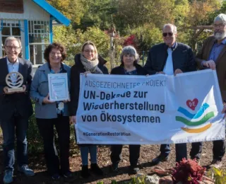 Sechs Personen stehen nebeneinander im Freien, zwei halten eine Auszeichnung und eine Urkunde, vier halten ein Banner mit der Aufschrift 'Ausgezeichnetes Projekt - UN-Dekade zur Wiederherstellung von Ökosystemen. #GenerationRestoration', im Hintergrund Gartenhäuschen und Bäume. 