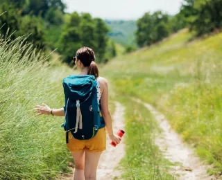 Eine Frau mit einem Rucksack auf dem Rücken geht einen Wanderweg entlang. Sie trägt eine Sonnenbrille und mit ihrer linken Hand streift sie die Gräser am Wegesrand, in ihrer rechten Hand hält sie eine rote Trinkflasche.